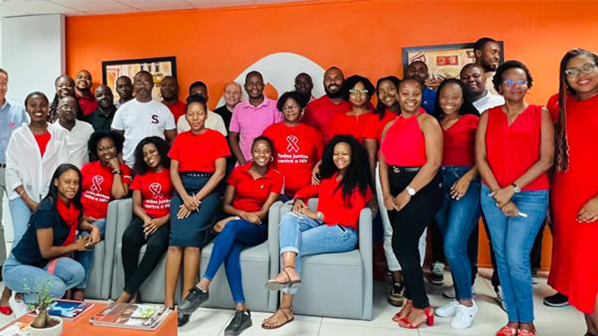 Fotografia de várias pessoas dentro de uma sala. Algumas em pé, outras sentadas em sofás cinzentos. Vestem, maioritariamente, camisolas de cor vermelha. A parede de fundo é cor-de-laranja e contém o logótipo da Galp a branco.