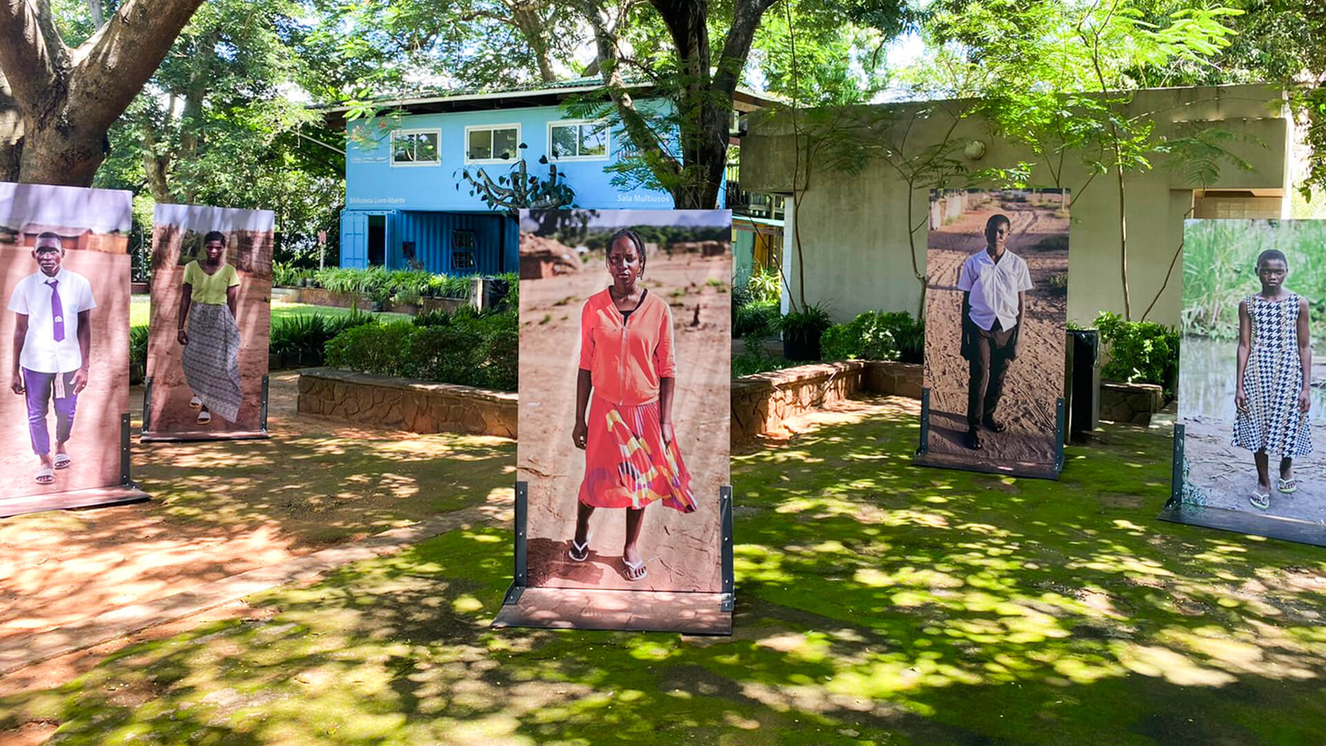 Fotografia de um jardim, com dois edifícios ao fundo; à esquerda, um azul e à direita um amarelo. No jardim, vê-se cinco fotografias de pessoas, em ponto grande, expostas.