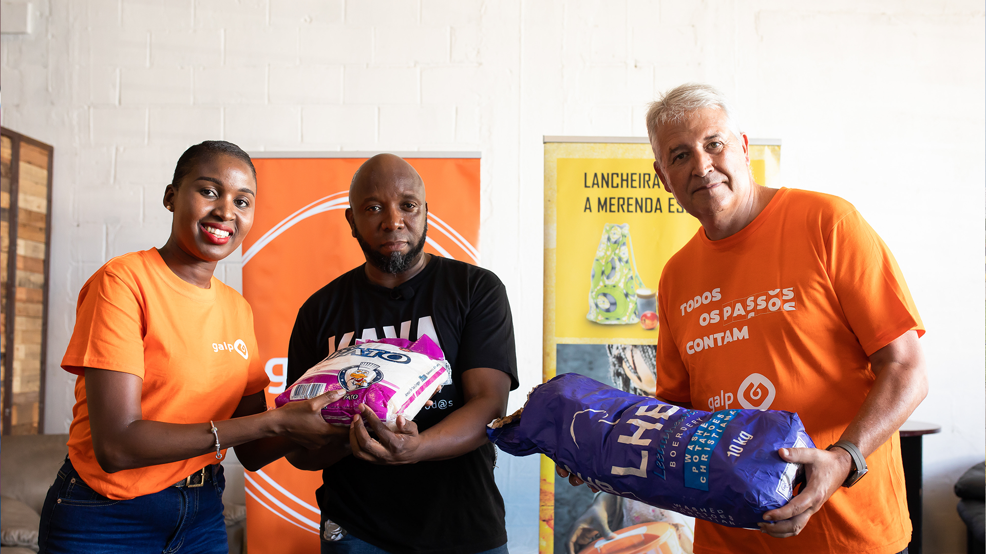 Fotografia de três pessoas, duas delas com uma tshirt cor-de-laranja com o logótipo da Galp. Seguram dois sacos com materiais.