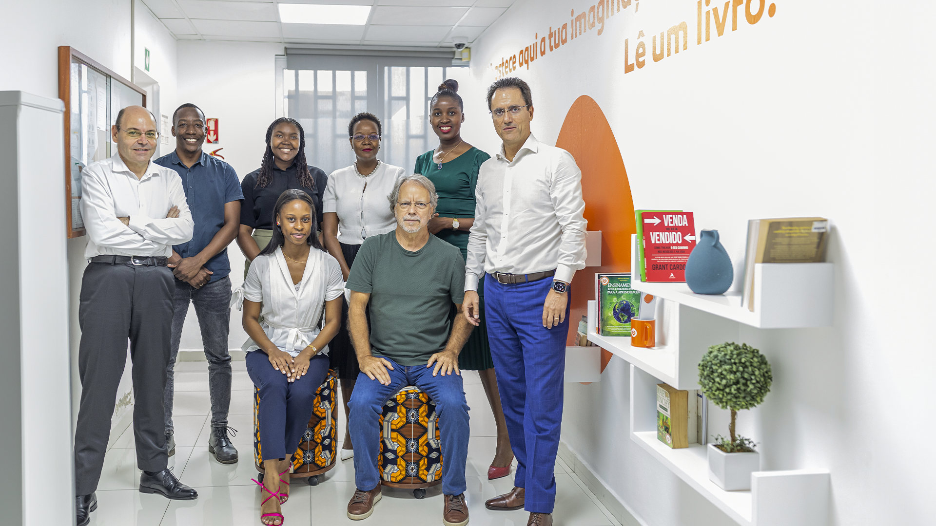 Fotografia de várias pessoas dentro de uma sala. Algumas estão em pá e duas estão sentadas em bancos. 