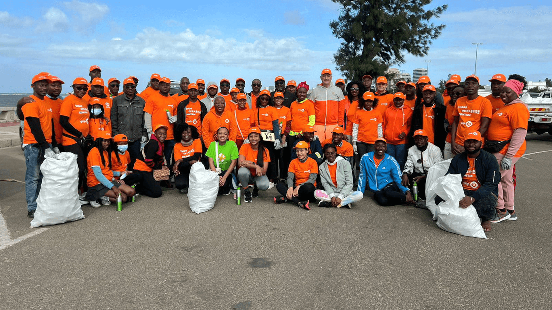 Fotografia ao ar-livre de várias pessoas. Algumas estão em pé e outras ajoelhadas ou sentadas. Todas vestem tshirts cor-de-laranja com o logótipo da Galp inscrito a branco. A grande maioria também utiliza um boné cor-de-laranja com o logóttipo da Galp a branco.
