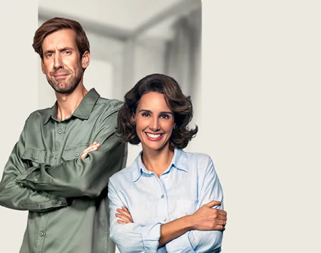 Fotografia de um casal, lado-a-lado, com os ombros encostados um ao outro. Estão em pé. Ele, do lado esquerdo, veste uma camisa verde e umas calças de ganga. Ela, do lado direito, veste uma camisa azul-clara e umas calças beges. São ambos morenos. O fundo da imagem é bege.