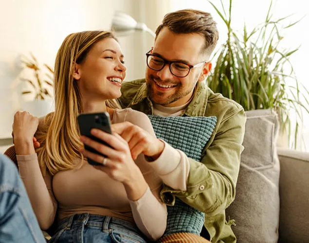 Fotografia de um casal sentado no sofá, dentro de casa. A senhora está sentada à frente do senhor, encostada a ele. Ela segura um telemóvel enquanto olha para ele, a rir. Ele está a olhar para o telemóvel e a apontar para o mesmo.