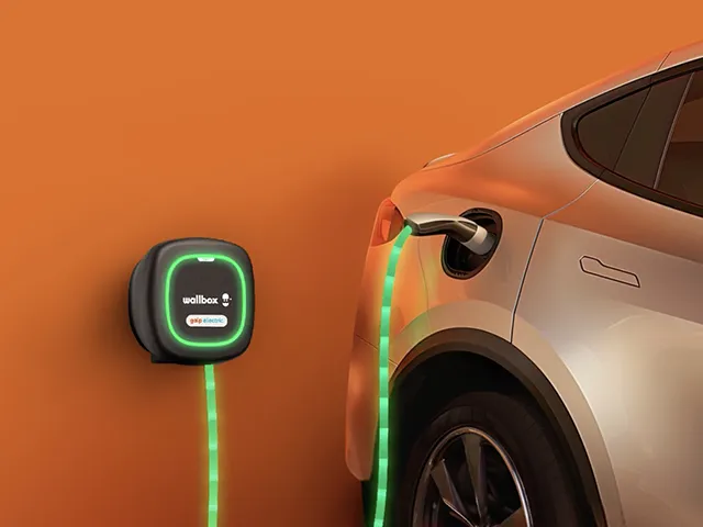 Carregador eléctrico de parede ligado a um automóvel eléctrico, com cabo iluminado a verde, num ambiente residencial com fundo laranja.