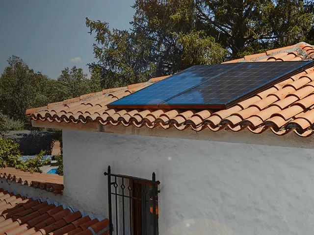 Painéis solares instalados no telhado de uma casa com telhas cerâmicas, integrados numa habitação rodeada de árvores, representando produção de energia solar.