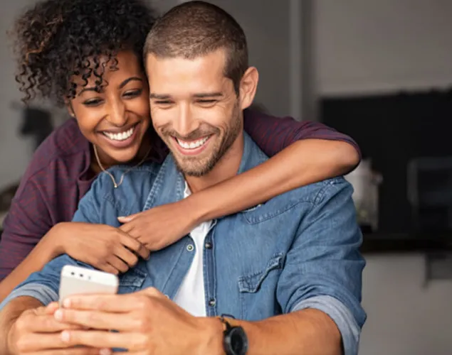 Casal dentro de casa. O homem está sentado numa cadeira a agarrar no telemóvel com ambas as mãos. Atrás dele, uma senhora a abraçá-lo.
