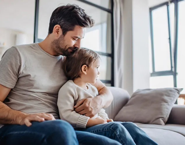 Pai sentado num sofá cinzento a abraçar uma criança numa sala luminosa, com plantas em primeiro plano e estantes ao fundo.