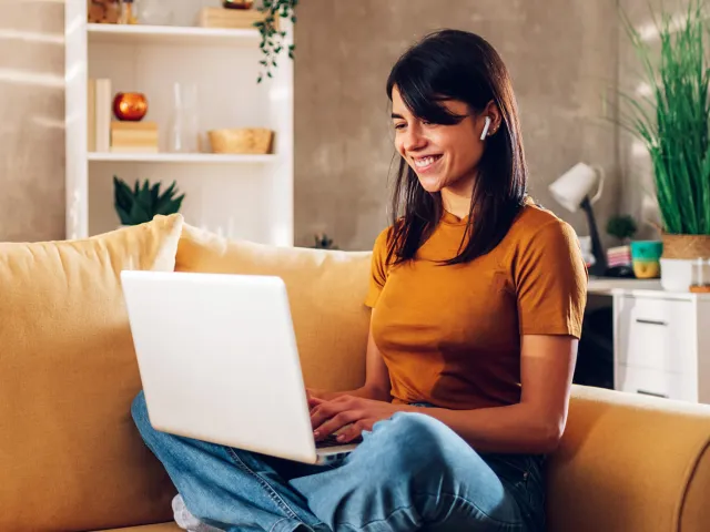 Uma mulher sorridente sentada num sofá amarelo, a usar um portátil com auriculares, num ambiente de sala de estar.