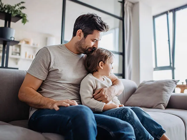 Pai sentado num sofá cinzento a abraçar uma criança numa sala luminosa, com plantas em primeiro plano e estantes ao fundo.