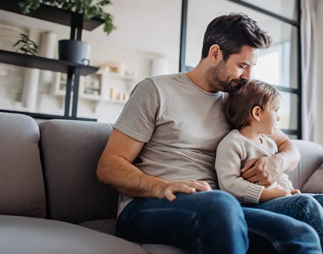 Pai sentado num sofá cinzento a abraçar uma criança numa sala luminosa, com plantas em primeiro plano e estantes ao fundo.
