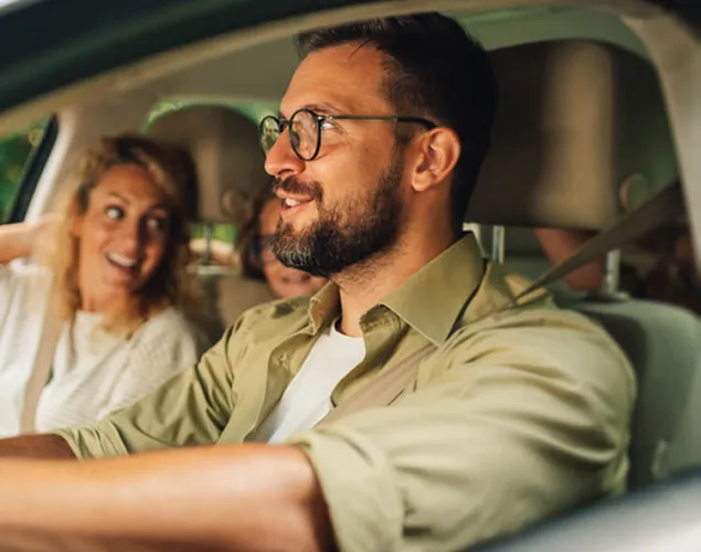 Fotografia de uma família dentro de um carro. Vê-se um homem ao telefone, veste uma camisa bege e usa óculos. Ao lado, uma senhora de cabelo ondulado, veste uma camisola branca. Atrás, vê-se uma criança desfocada.