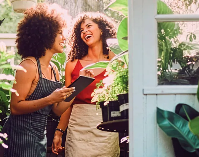 Fotografia de duas senhoras a rir numa marquise de uma casa. Tanto a marquise quanto a entrada para a mesma contém várias plantas verdes. As senhoras estão ambas de avental.