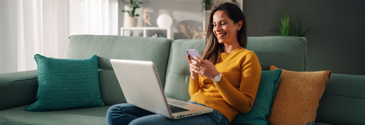 Uma mulher sorridente sentada num sofá verde, a usar o telemóvel e com um portátil no colo, num ambiente de sala de estar.