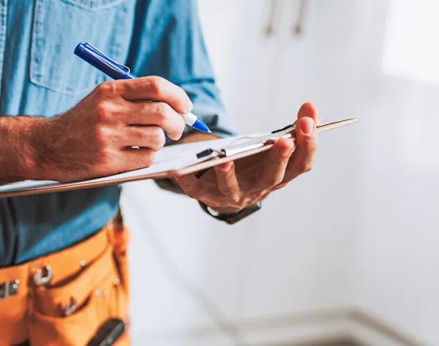 Fotografia meio corpo de um homem. Tem uma camisa de ganga e umas calças pretas. Tem um cinto de ferramentas cor-de-laranja. Segura uma caneta e uma folha que está a preencher.