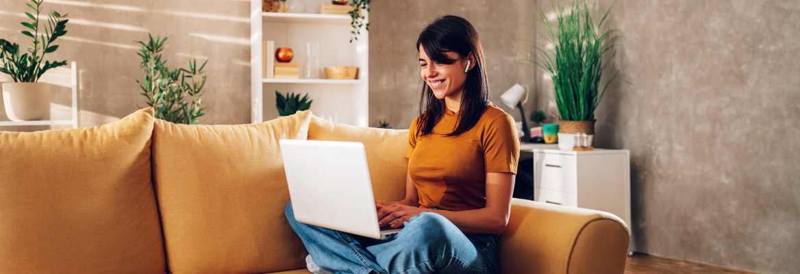 Uma mulher sorridente sentada num sofá amarelo, a usar um portátil com auriculares, num ambiente de sala de estar.