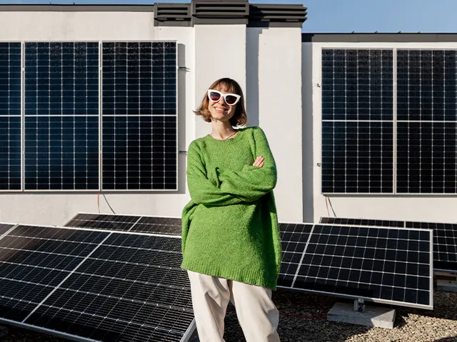 Mulher de braços cruzados num telhado com painéis solares, a sorrir para a câmara, representando um produtor de energia solar.