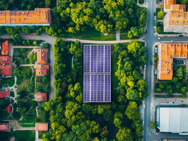 Vista aérea de uma cidade com grandes painéis solares azuis no centro, rodeados por densas áreas verdes e edifícios com telhados laranja, com estradas e carros visíveis.