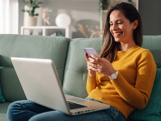 Uma mulher sorridente sentada num sofá verde, a usar o telemóvel e com um portátil no colo, num ambiente de sala de estar.