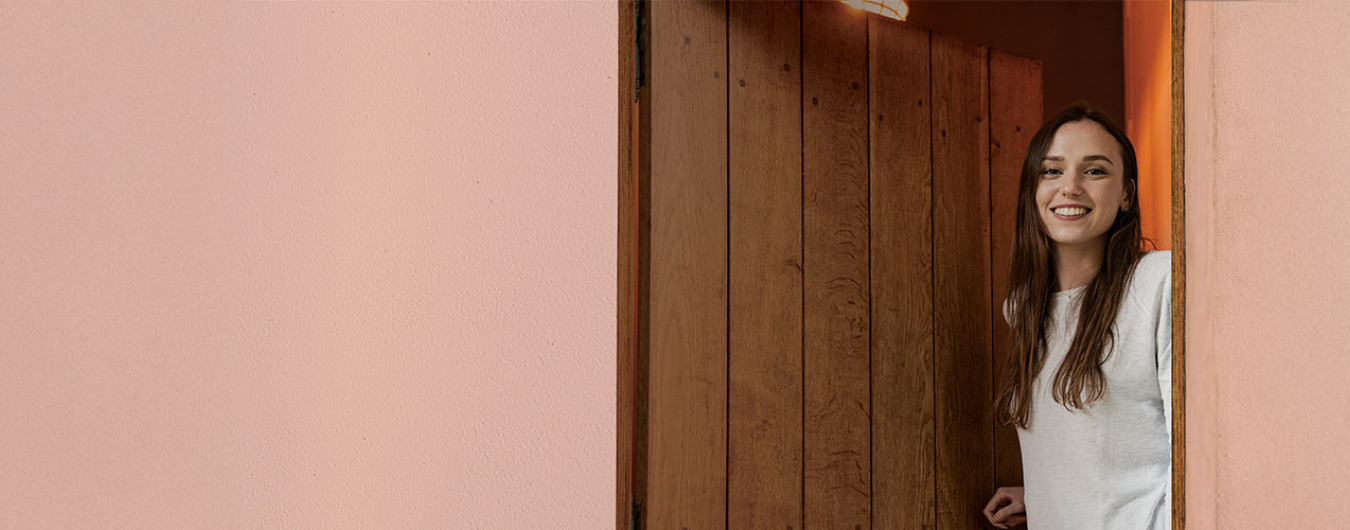 Uma mulher sorridente, de cabelos castanhos e camisola clara, espreita por detrás de uma porta de madeira, ladeada por paredes cor-de-rosa.