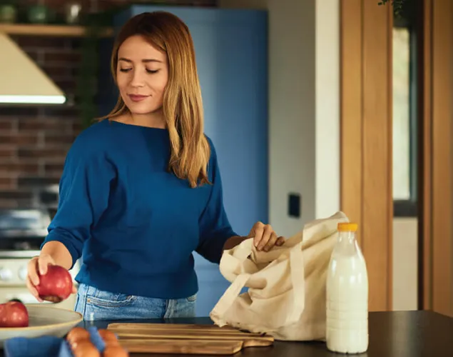 Fotografia de uma senhora numa cozinha a tirar maças de um saco de compras bege. O saco, bem como o prato onde está a colocar as maçãs estão em cima de uma bancada.