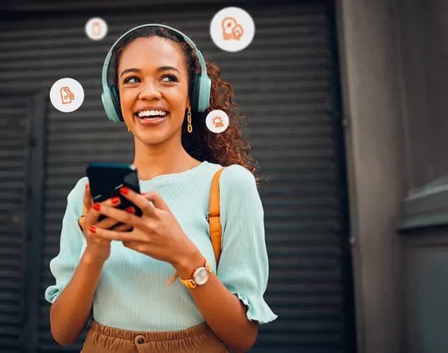 Senhora na rua a segurar o telemóvel com ambas as mãos. Está a sorrir. Veste uma camisola azul-clara e umas calças castanhas. Está de fones.