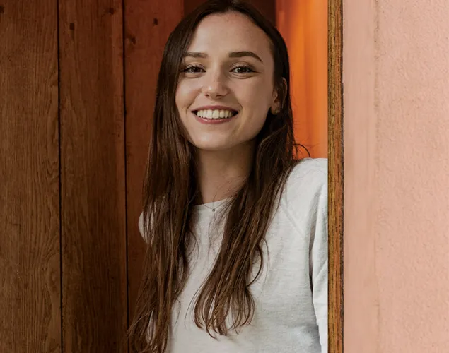 Uma mulher sorridente, de cabelos castanhos e camisola clara, espreita por detrás de uma porta de madeira, ladeada por paredes cor-de-rosa.