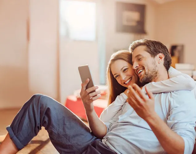 Casal sentado no chão de casa a sorrir enquanto consulta um smartphone. A imagem transmite conforto no lar, gestão de serviços domésticos e soluções de energia para casa.