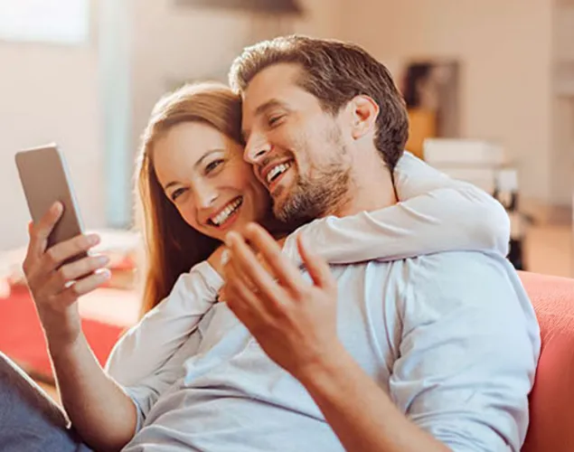 Casal sentado no chão de casa a sorrir enquanto consulta um smartphone. A imagem transmite conforto no lar, gestão de serviços domésticos e soluções de energia para casa.