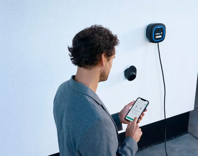 Fotografia de um omem de costas, vê-se que está a agarrar num telemóvel com as duas mãos. À esquerda, uma parece com um carregador elétrico Galp Wallbox na mesma.