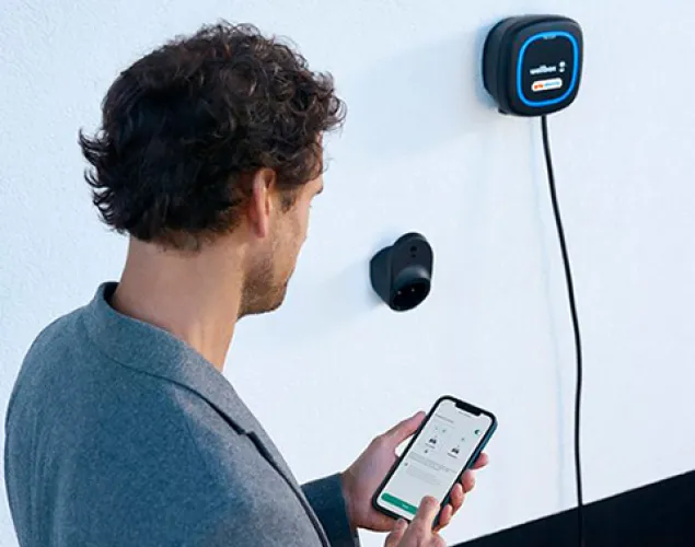 Fotografia de um omem de costas, vê-se que está a agarrar num telemóvel com as duas mãos. À esquerda, uma parece com um carregador elétrico Galp Wallbox na mesma.